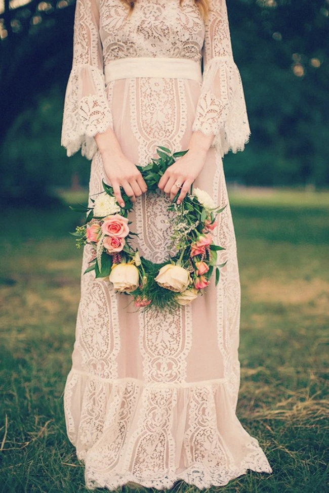 Mulher usando um vestido de noiva inspirado nos anos 70. Ela segura uma grinalda de flores com ambas as mãos em frente ao corpo. Ela está em um amibiente ao ar livre com grama. A foto só mostra o corpo da mulher.