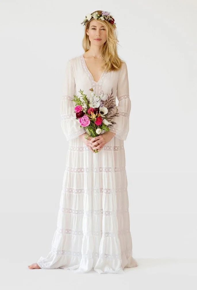 Mulher loira usando um vestido de noiva branco inspirado dos anos 70. Ela está segurando um buque de flores coloridas com ambas as mãos em frente ao corpo e olha diretamente para a câmera.