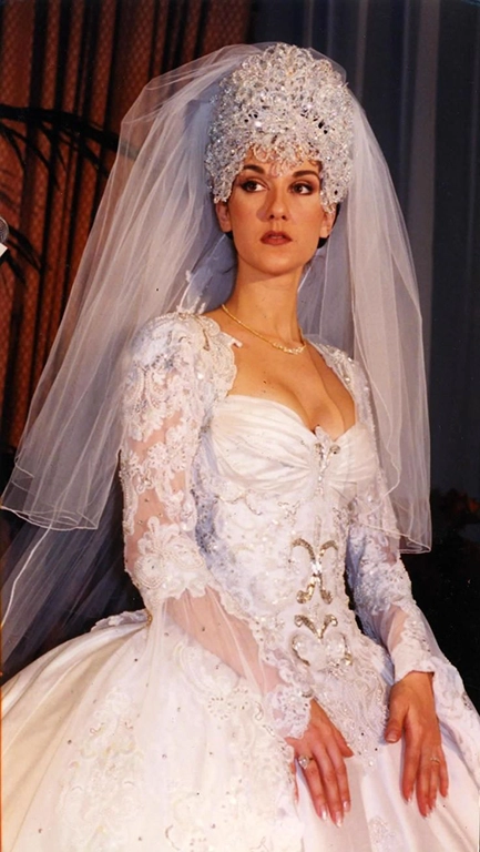 Mulher de cabelos castanho escuro olhando para longe. Ela usa um vestido de noiva dos anos 80 e uma coroa brilhantee um véu na cabeça.
