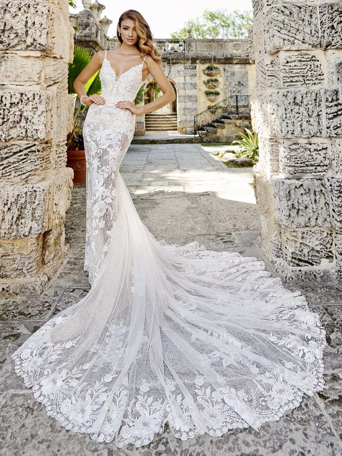 Mulher usando um vestido de noiva branco, modelo sereia com cauda longa. Ela faz uma pose para a foto com as mãos na cintura.