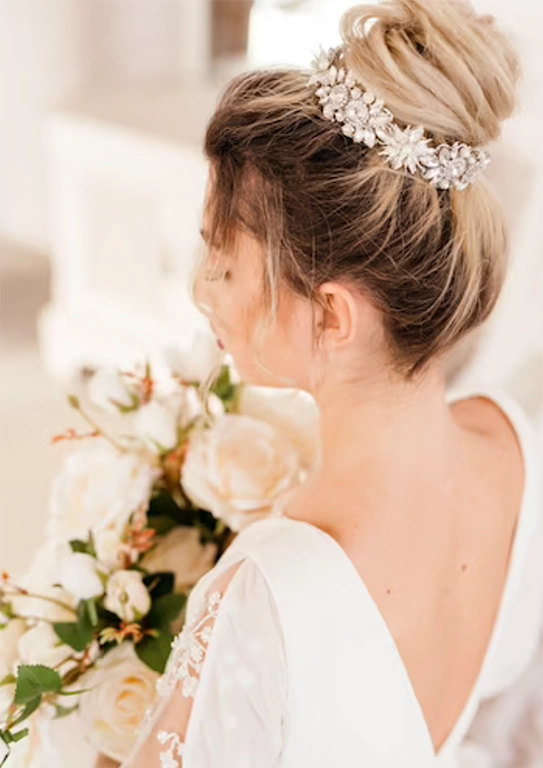 Mulher de cabelos castanhos usando um vestido de noiva e uma grinalda prata em seus cabelos em coque. Ela está de costas e segura um buque de flores em tons claros com ambas as mãos.