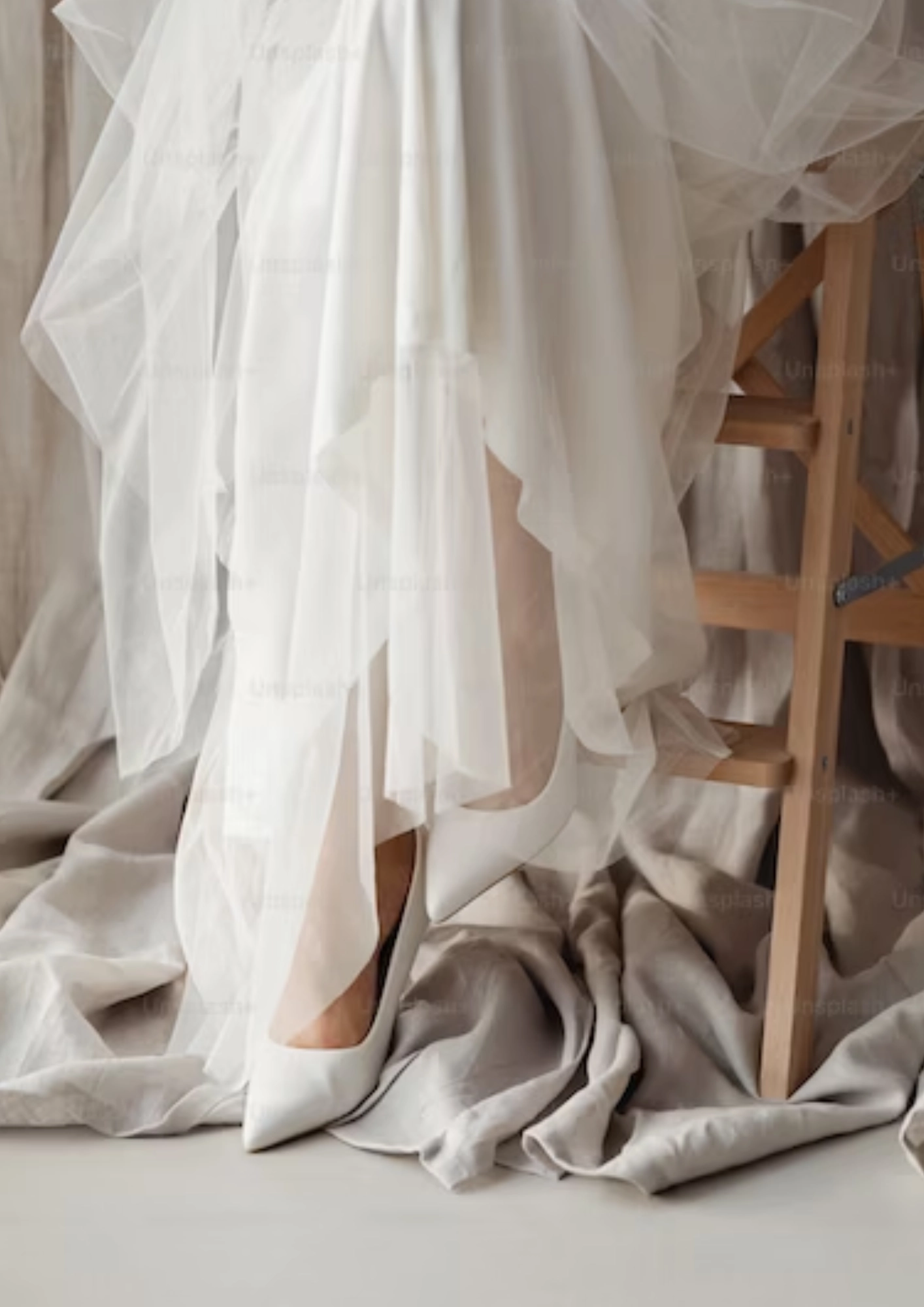 Mulher sentada em um banquino de madeira. A foto foca nos sapatos brancos e na barra do vestido de noiva. No chão, podemos ver um tecido beje.