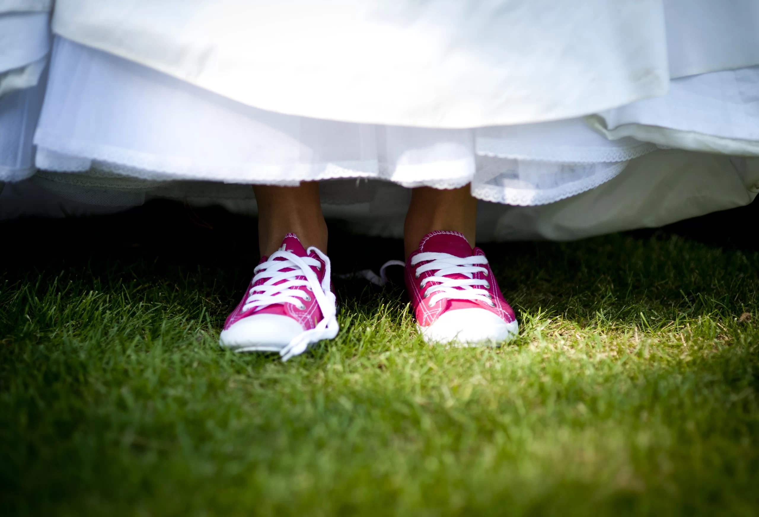 Mulher usando tênis na cor rosa e um vestido de noiva branco em cima da grama. A foto mostra um pouco da grama, os sapatos e a barra do vestido da mulher.