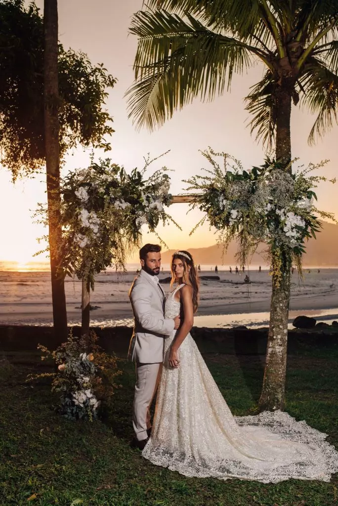 Casal em uma cerimônia de casamento na praia ap por do sol. A noiva usa um vestido de noiva no tom off white.