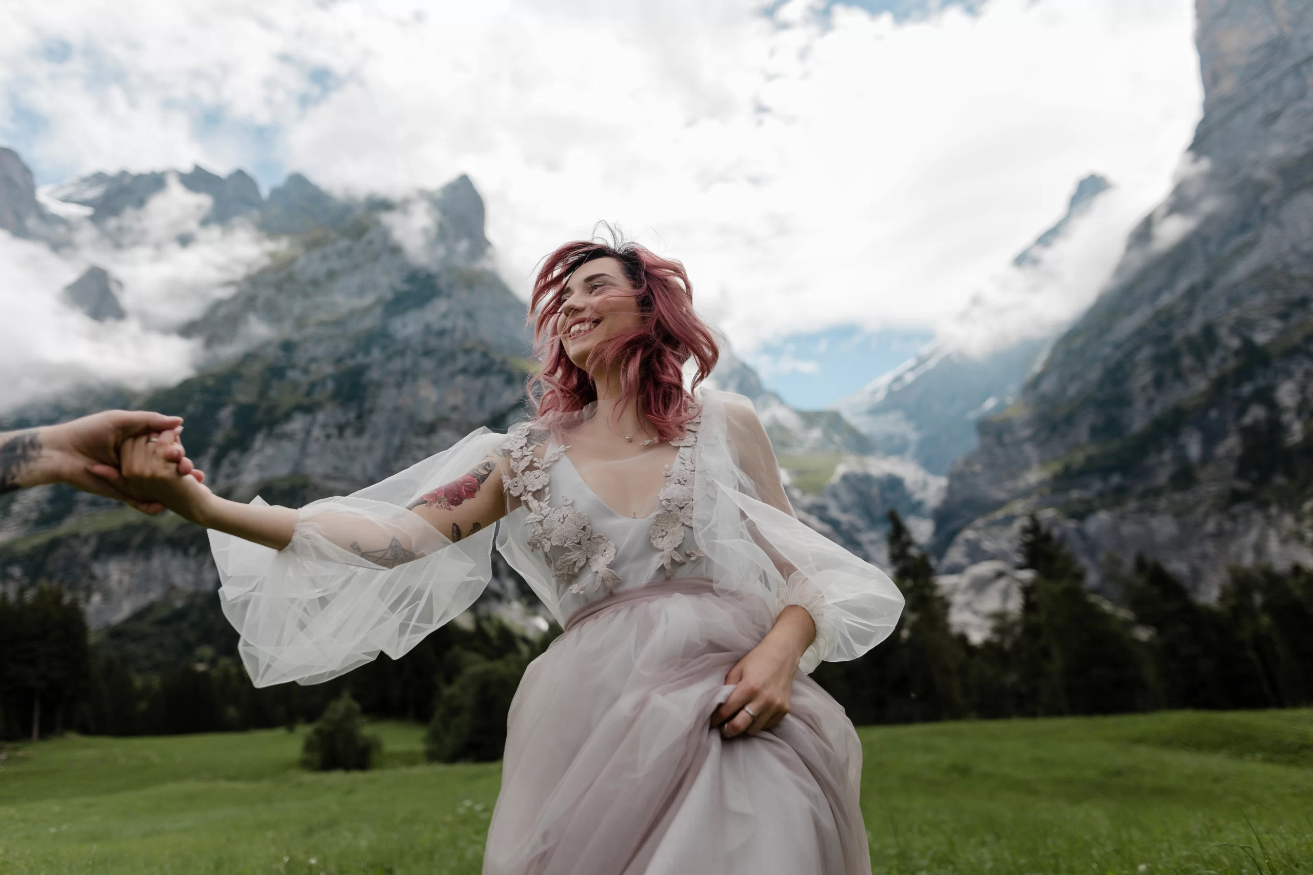 Mulher ruiva feliz correndo com um vestido de noiva. Ela está na grama ao ar livre com montanhas e arvores aparecendo ao fundo da imagem.