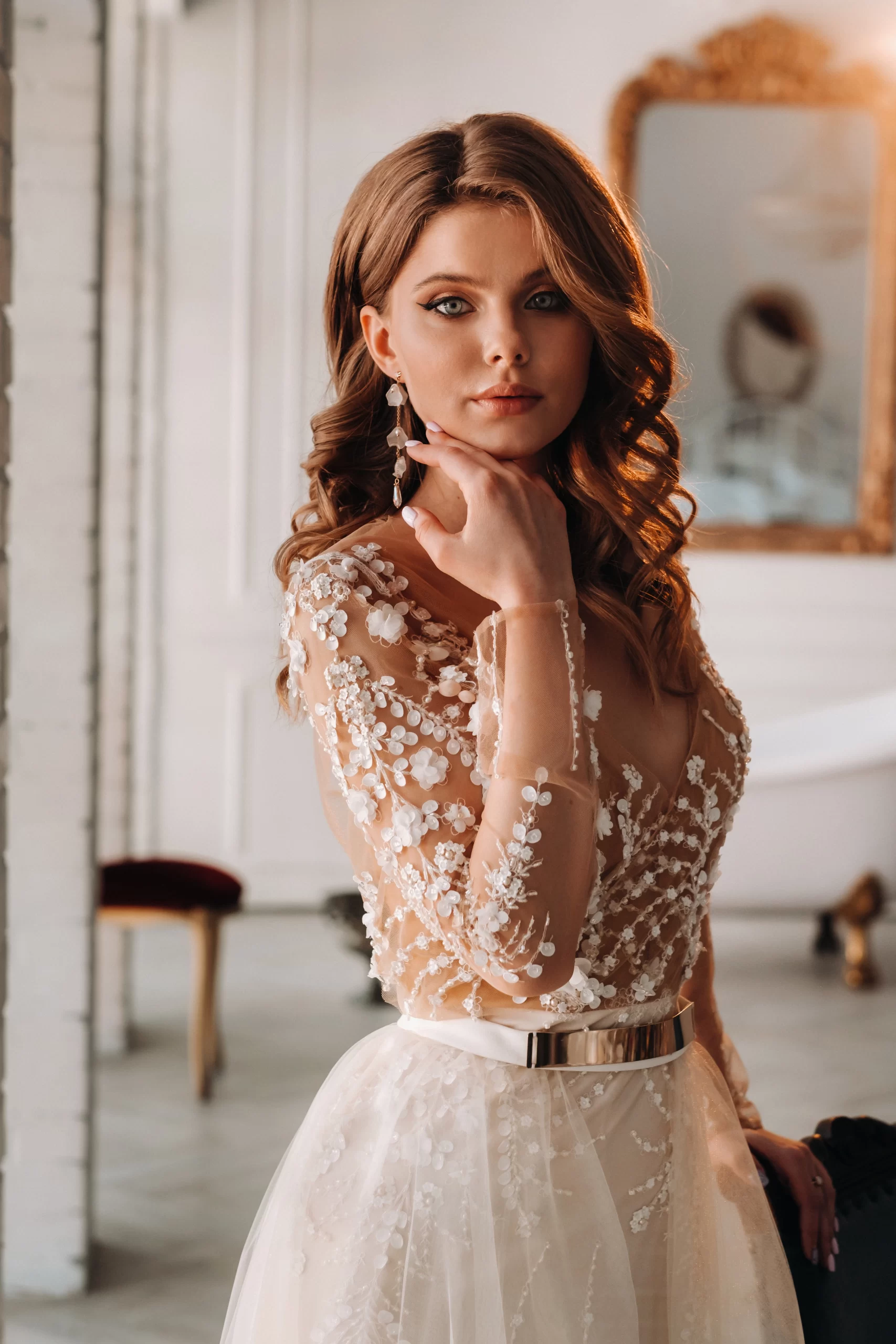 Mulher ruiva usando um vestido de noiva no tom rose com detalhes brancos. Ela está em uma sala e faz uma pose com a mão no rosto enquanto olha para a cãmera.
