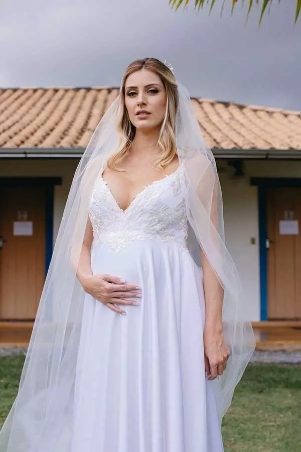 Mulher lusando um vestido de noiva soltinho com um decote em V. Ela olha para a câmera e segura a barriga com uma das mãos.