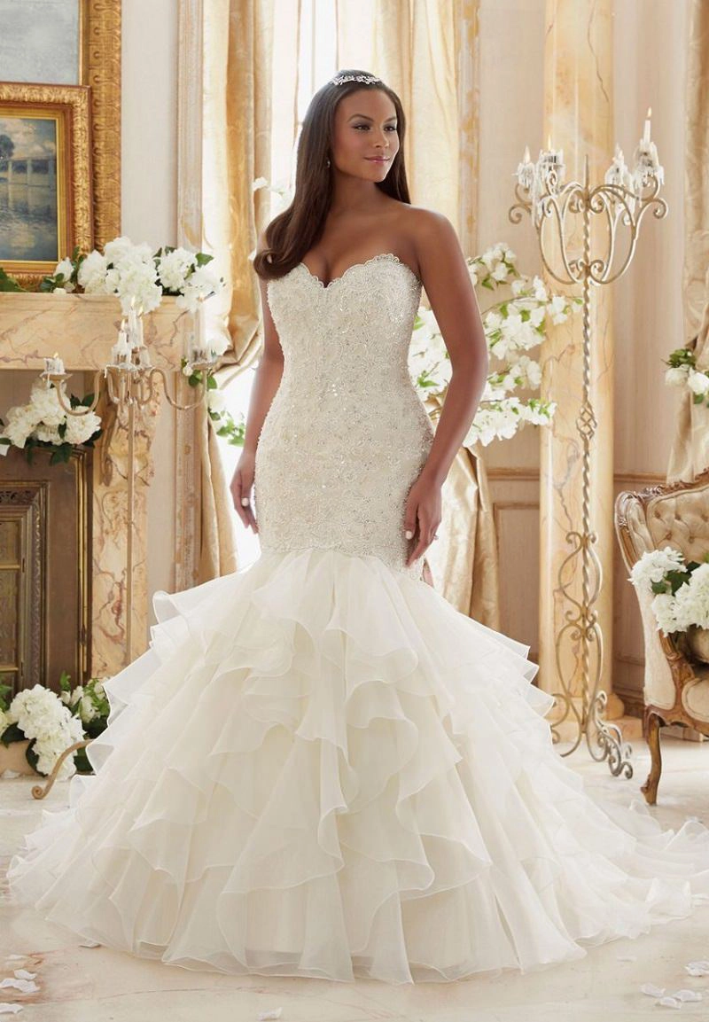 Mulher de cabelos pretos usando um vestido de noiva sereia branco com detalhe da cauda voal. Ela está em uma sala decorada com flores brancas. A mulher olha para o lado e sorri levemente.