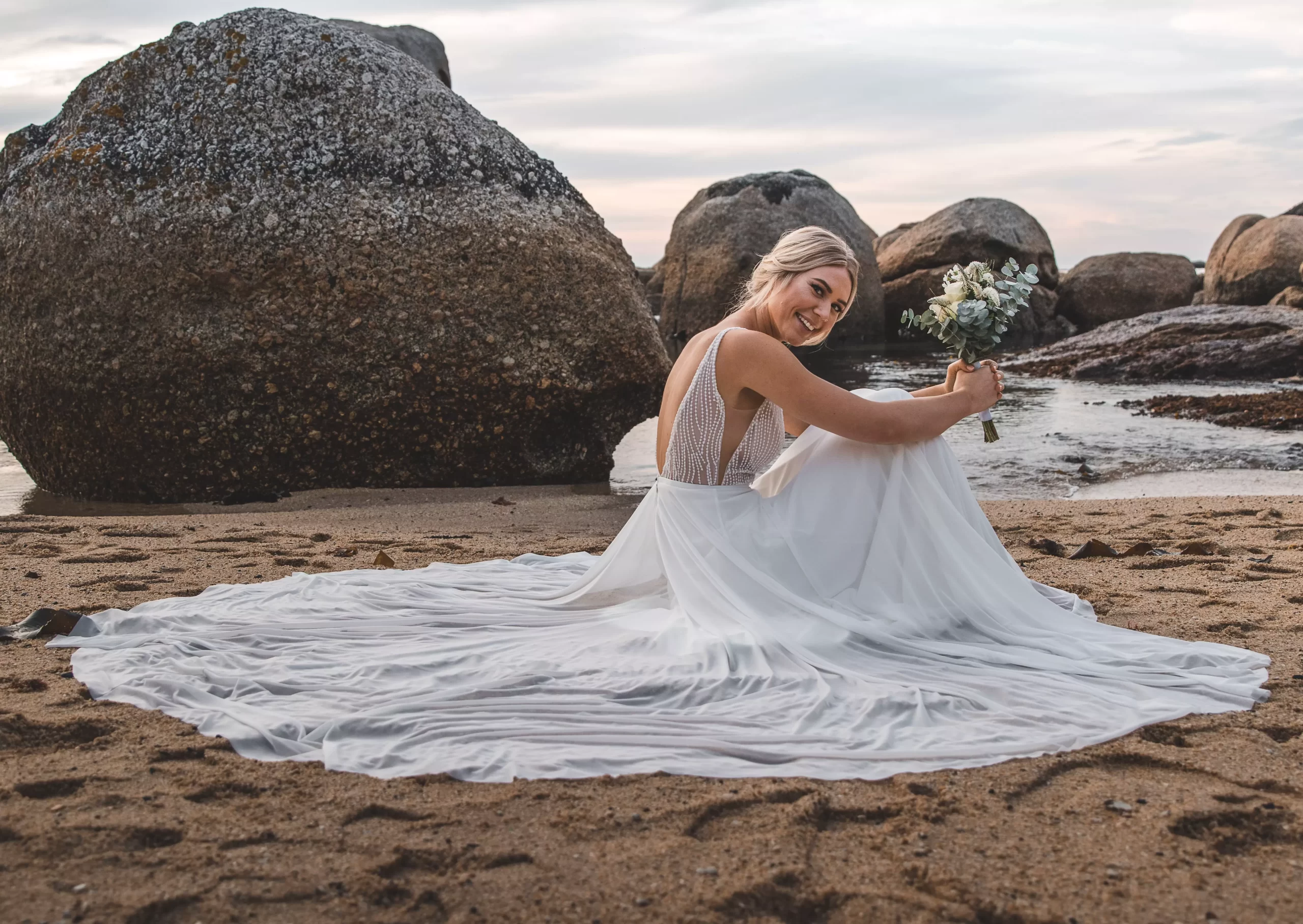 Mulher usando vestido de noiva simples na praia. Ela está sentada na areia e sori para a camera segurando um buque de flores.