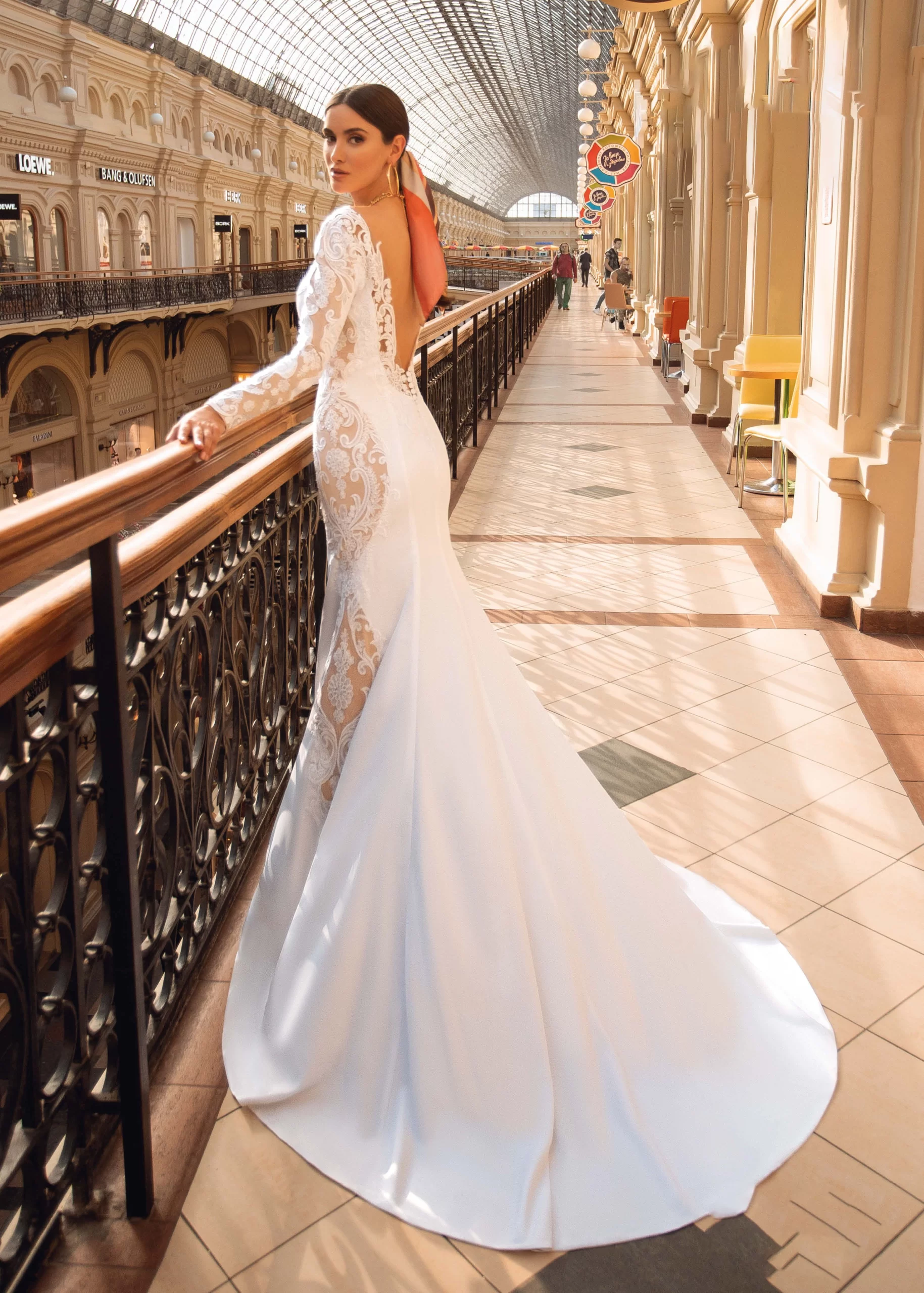 Mulher de cabelos castanhos em um shopping usando um vestido de noiva sereia branco transparente. Ela está segurando a grade e olhando para a câmera.