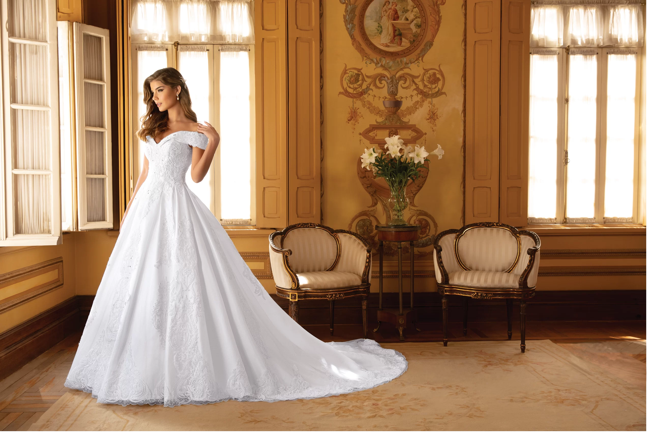 Mulher de cabelos castanhos em uma sala usando um vestido de noiva branco. Ela está de lado e olha para longe  com uma das mãos no ombro.