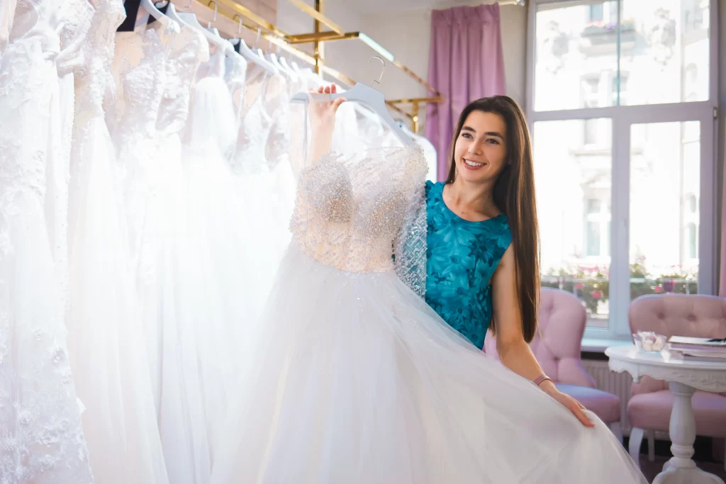 Mulher de cabelos castanhos e blusa azul em uma loja de vestidos de noivas. Ela segura um vestido e sorri olhando para longe.