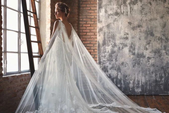 Mulher usando um vestido de noiva branco com uma capa transparente nas costas. A mulher está olhando pela janela.