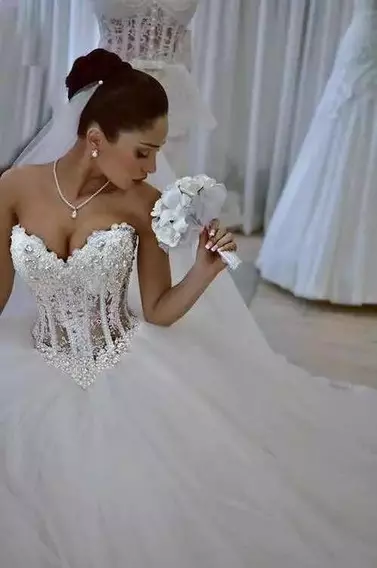 Mulher usando um vestido de noiva branco com corpete. A mulher está fazendo uma pose segurando um buque de flores com uma das mãos.