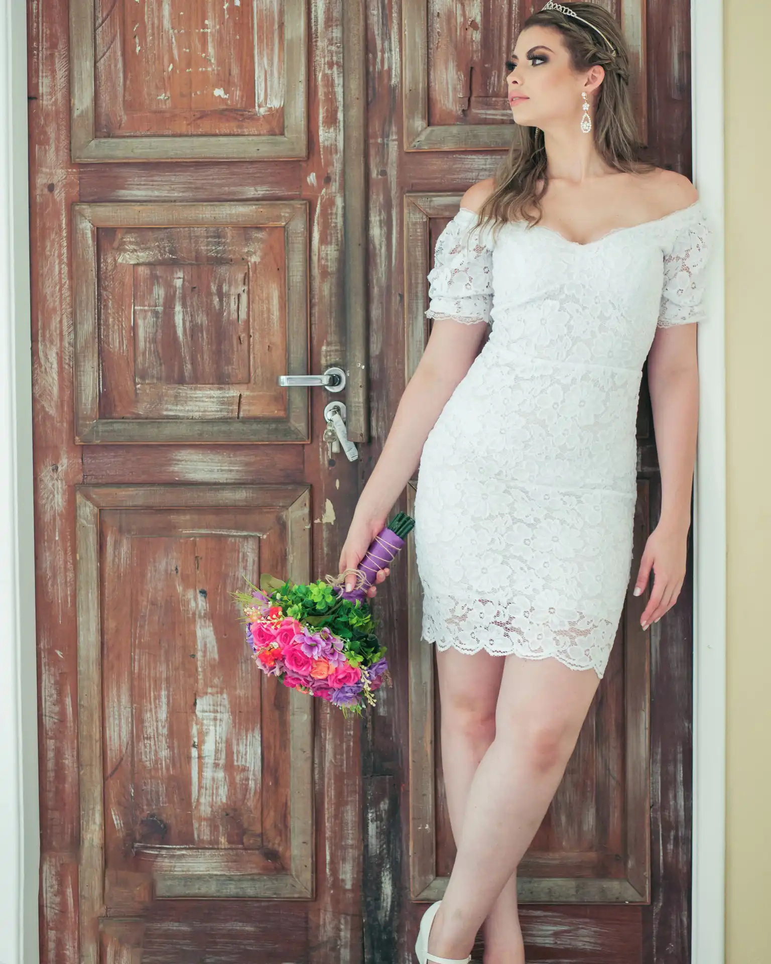 Mulher usando um vestido de noiva civil branco. A mulher está recostada em uma porta e segura um buque de flores.