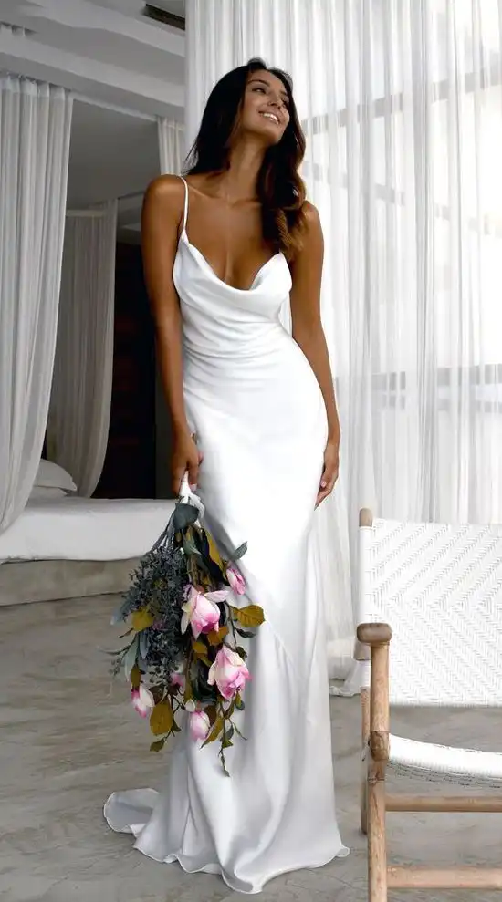 Mulher vestindo um vestido de noiva branco de modelo tubinho feito de seda. Ela segura um buque de flores em sua mão.