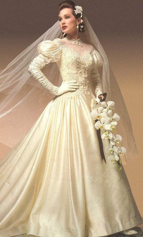 Mulher de cabelos castanho escuro usando um vestido de noiva na cor off white, inspirado nos anos 80. Ela faz uma pose com uma mão na cintura e olha para o lado. Em uma das mãos ela segura um ramo de flores brancas.