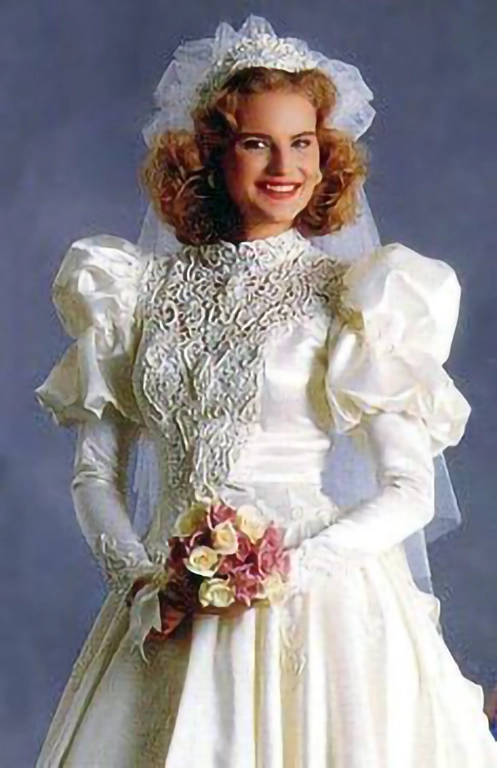 Mulher de cabelos castanho claro curto usando um vestido de noiva dos anos 80. Ela segura um buquê de flores com ambas as mãos nem frente ao corpo e sorri para a câmera.