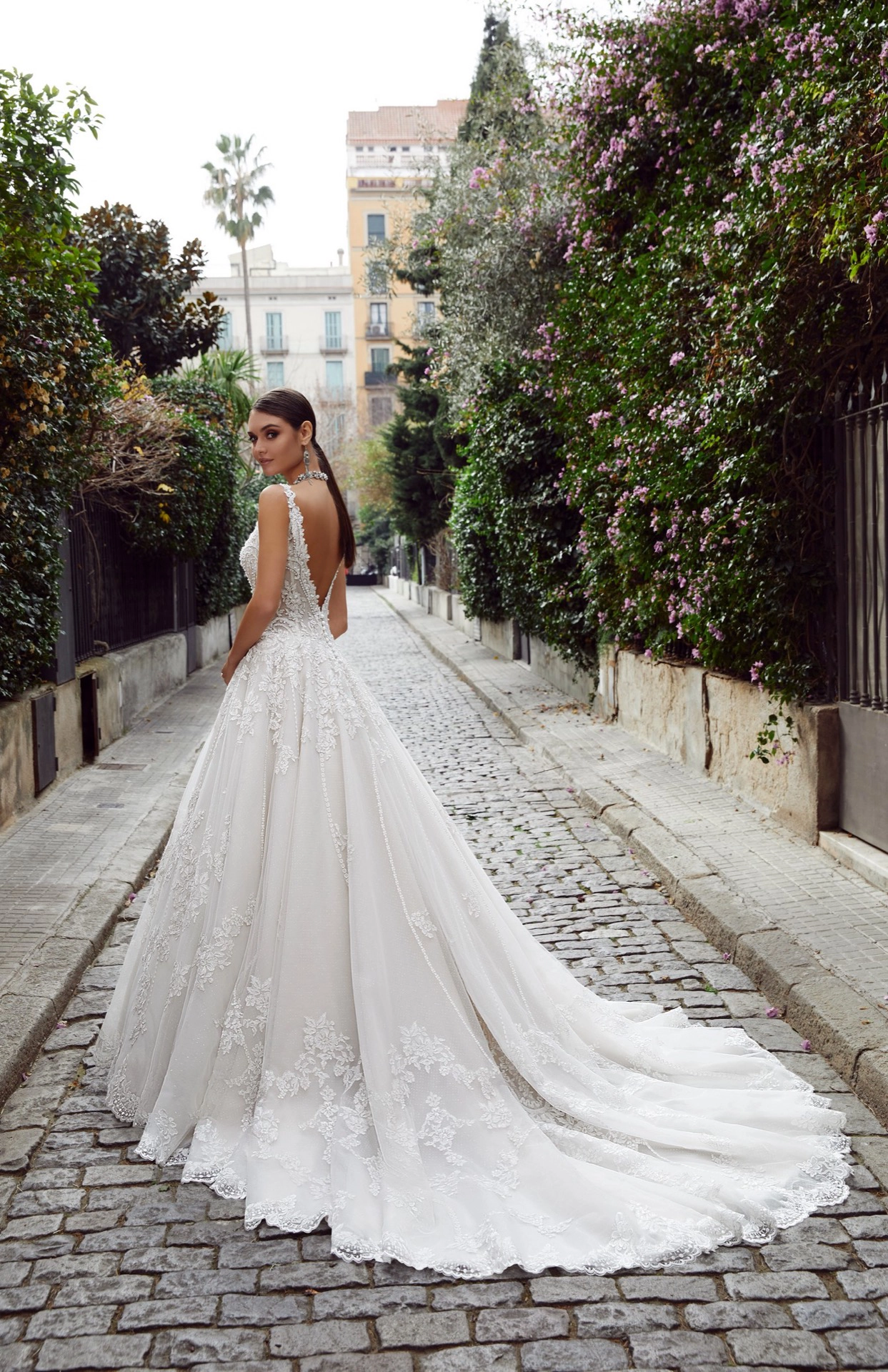 Mulher de cabelos castanho escuro usando um vestido d noiva branco. Ela está de costas e olha para trás, diretamente para a câmera. A mulher está no meio de uma ruazinha de paralelepipedos com vegetação em ambos os lados da ruazinha. Ao fundo vemos dois prédios antigos.