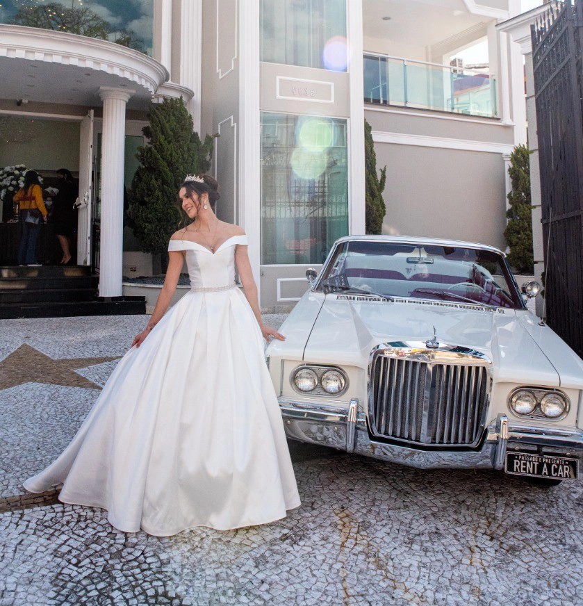 Mulher de cabelos castanhos usando um vestido de noiva branco estilo princesa. Ela está fazendo uma pose olhando para o lado e levemente para baixo. Ao lado da mulher há um carro. Ao fundo aparece uma mansão.