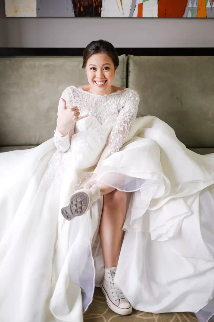 Mulher sentada em um sofá usando um vestido de noiva branco. Ela está usando um all-star nos pés e sorri para a câmera fazeno um joinha com uma das mãos.