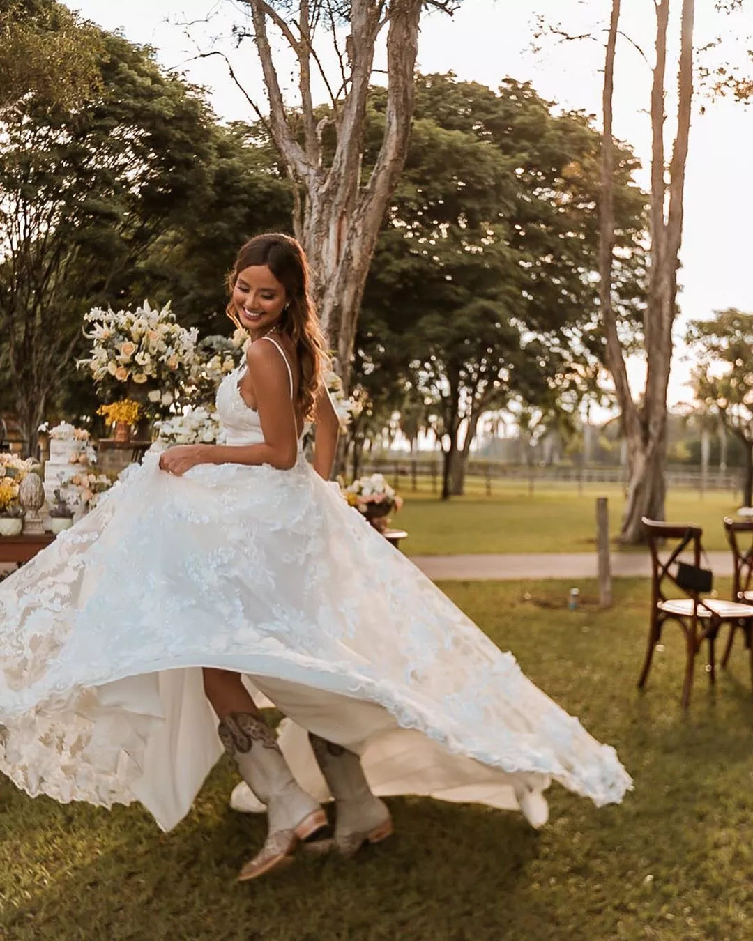 Mulher usando um vestido de noiva branco e bota. Ela está se movimentando de modo que o vestido rode e mostre as botas. Ela sorri e olha levemente para baixo.