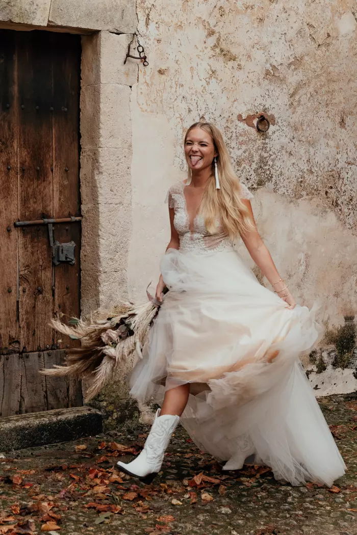 Mulher usando vestido de noiva branco com bota. Ela sorri para a câmera, puxa a barra do vestido com uma das mãos e com a outra segura um buque de flores.