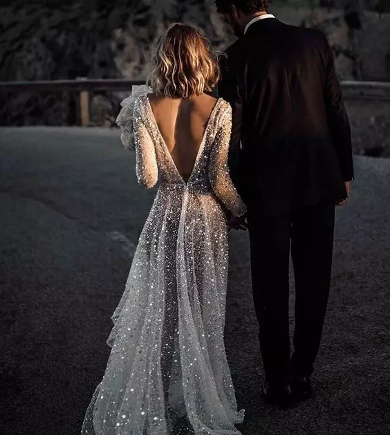 Mulher e homen de mãos dadas de costas andando para longe. A mulher usa um vestido de noiva branco com brilho e segura um buque. O homem está usando um terno preto.