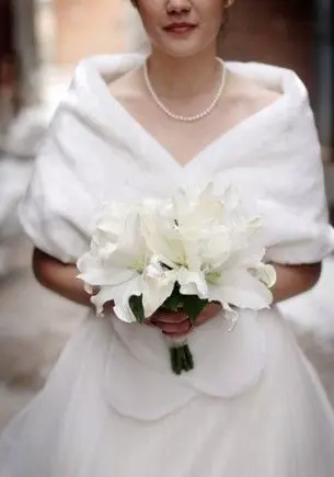 Mulher usando um vestido de noiva branco e um chale branco por cima. Ela segura um buque de flores com ambas as mãos.