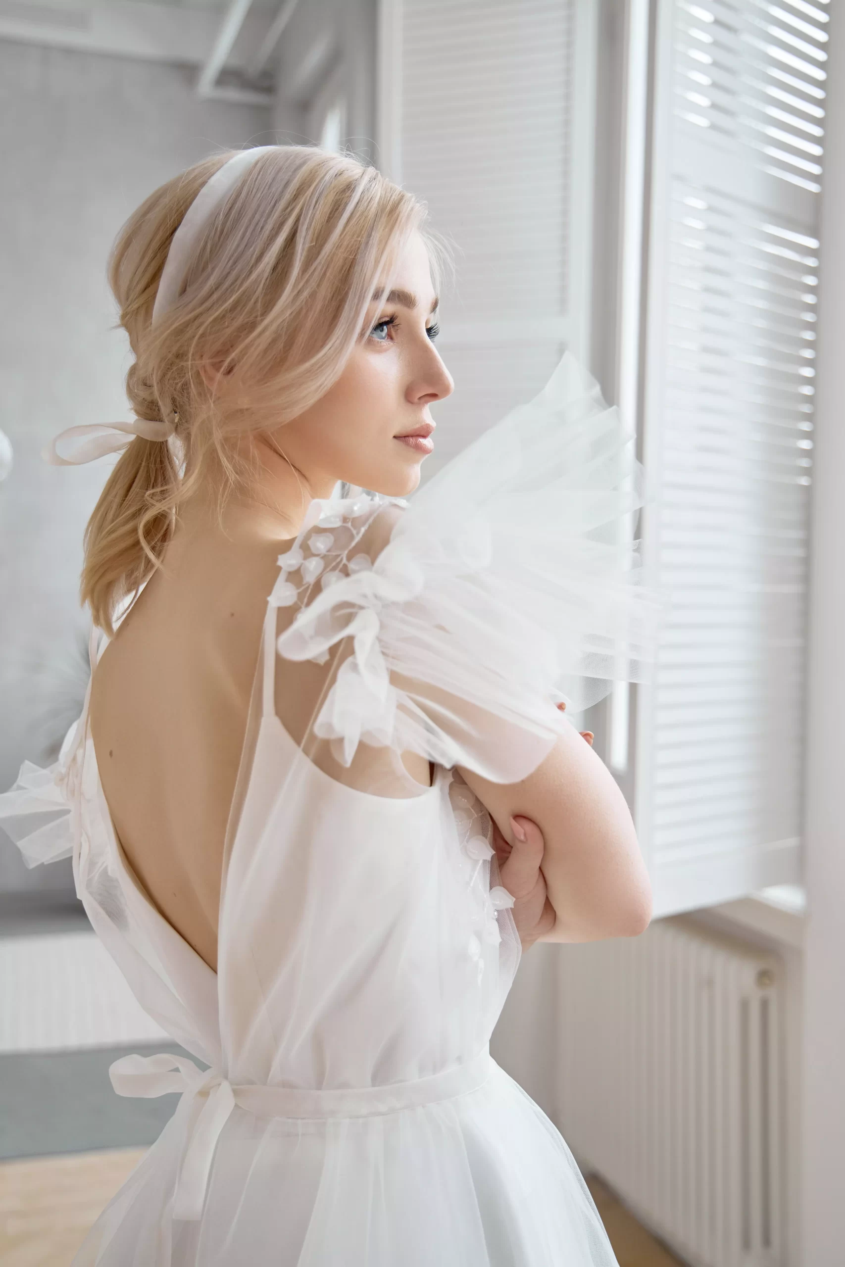 Mulher loira usando um vestido de noiva branco com mangas curtas de tule. Ela está com os braços cruzados e olha pela janela.