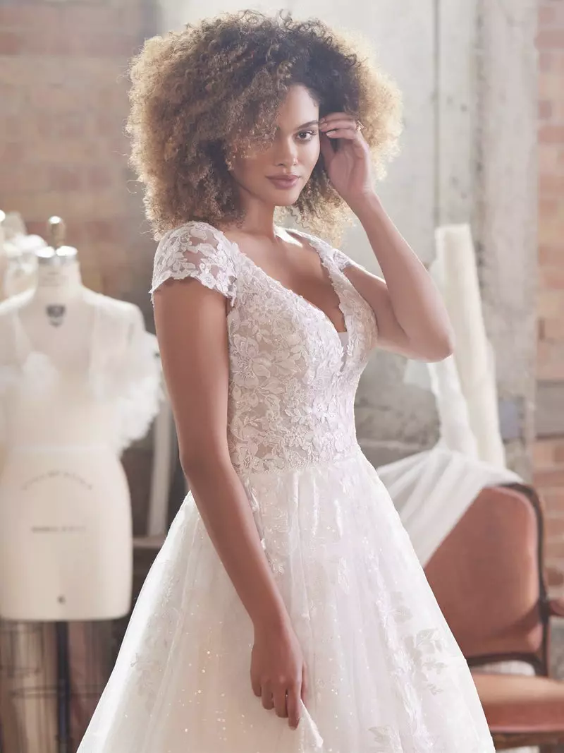 Mulher com o cabelo de cor castanho encaracolado usando um vestido de noiva branco. Ela está em um ateliê de roupas. A mulher faz uma pose com uma mão no cabelo e olha para a câmera,