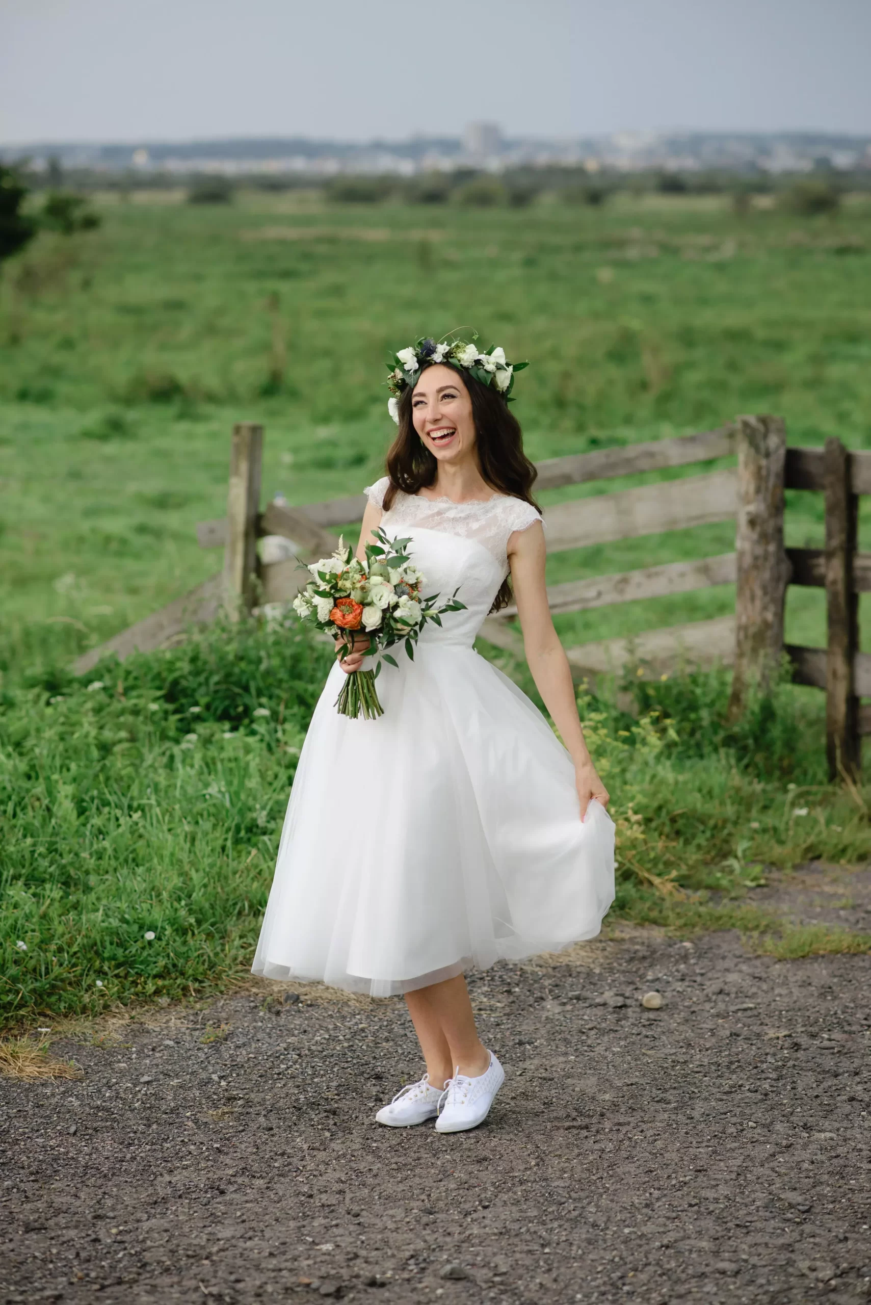 Mulher usando um vestido de noiva branco curto com manga cura. Ela sorri e olha para longe enquanto faz uma pose segurando a barra dos vestidos com uma das mãos enquanto segura um buque de flores com a outra mão.