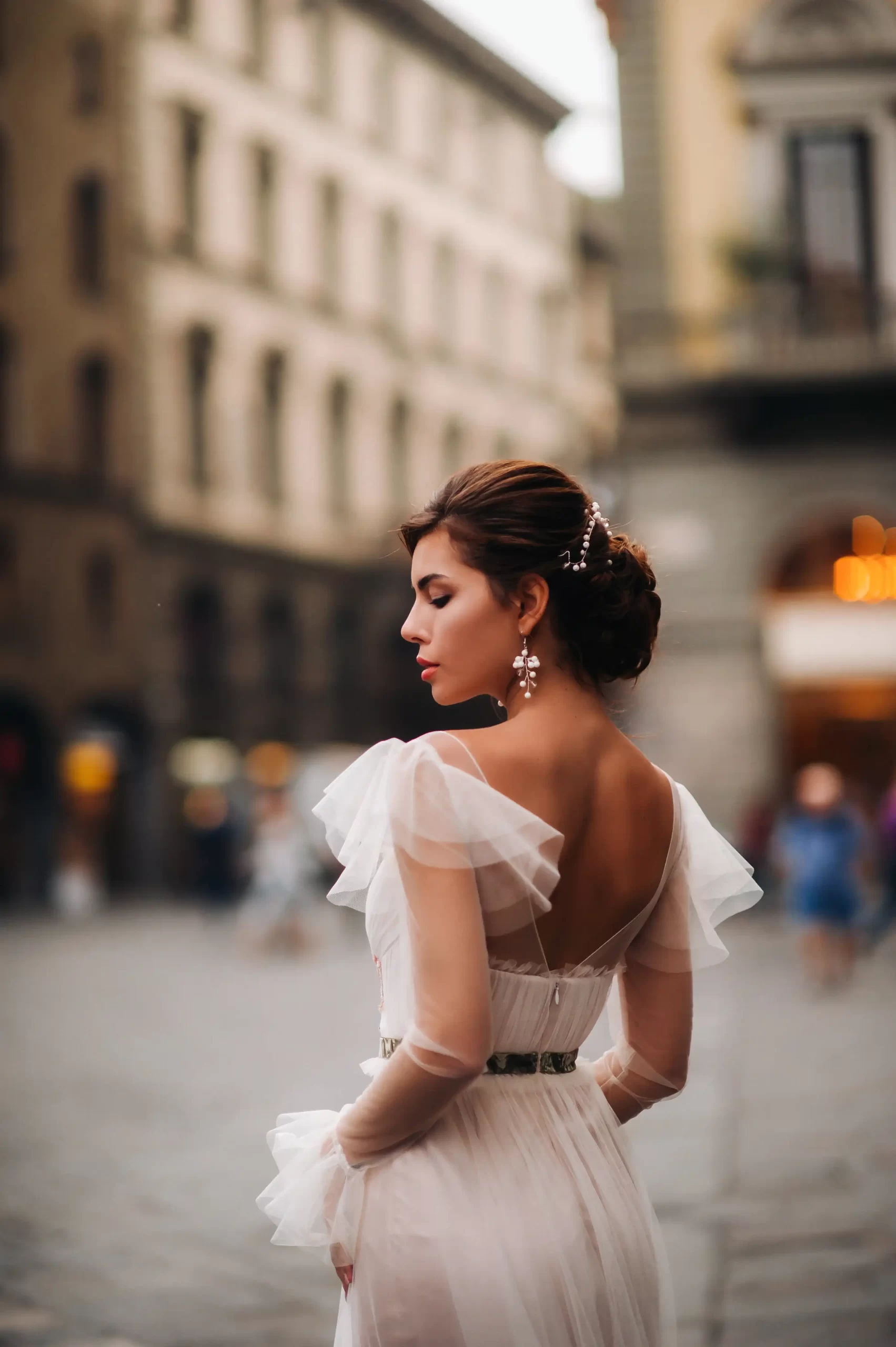 Mulher usando um vestido de noiva branco com mangas de tule. Ela esta de costas e olha para longe.