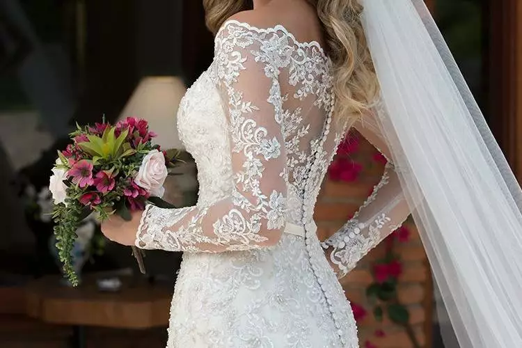 Mulher usando um vestido de noiva branco com mangas de renda longas. O foco da foto é na manga do vestido e a noiva segura um buque de flores em uma das mãos.