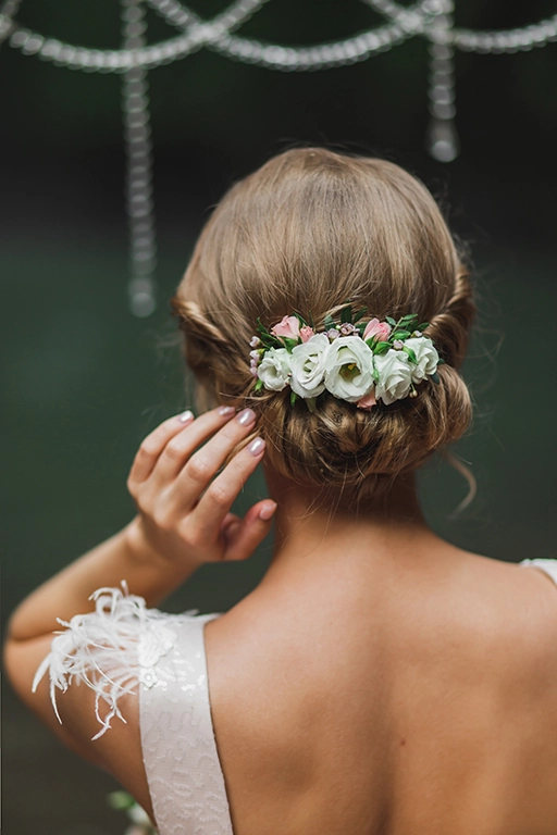 Mulher de costas com a mão no penteado dos cabelos. Ela tem cabelos castanho claro e seu cabelo está em um coque preso com um acessório de flores e folhas na cor branca, rosa e verde.