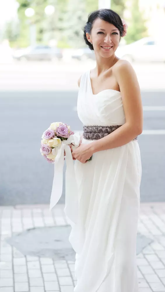 Mulher usando um vestido de noiva estilo grego. Ela segura um buque de flores nas mãos.