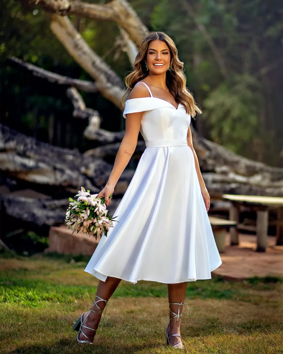 Mulher usando um vestido de noiva branco, evasê curto. A mulher sorri para a câmera e segura um buque de flores com uma das mãos.