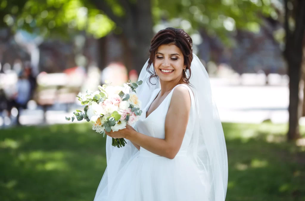 Mulher usando um vestido de noiva branco em um parque de dia. Ela sori para a camera e segura um buque de flores com as mãos.