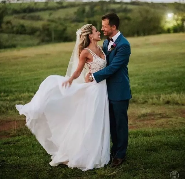 Mulher e homem em um ambiente campestre. Ele esta usando um terno e está abracando a mulher. Ela usa um vestido de noiva branco para casamento no campo.