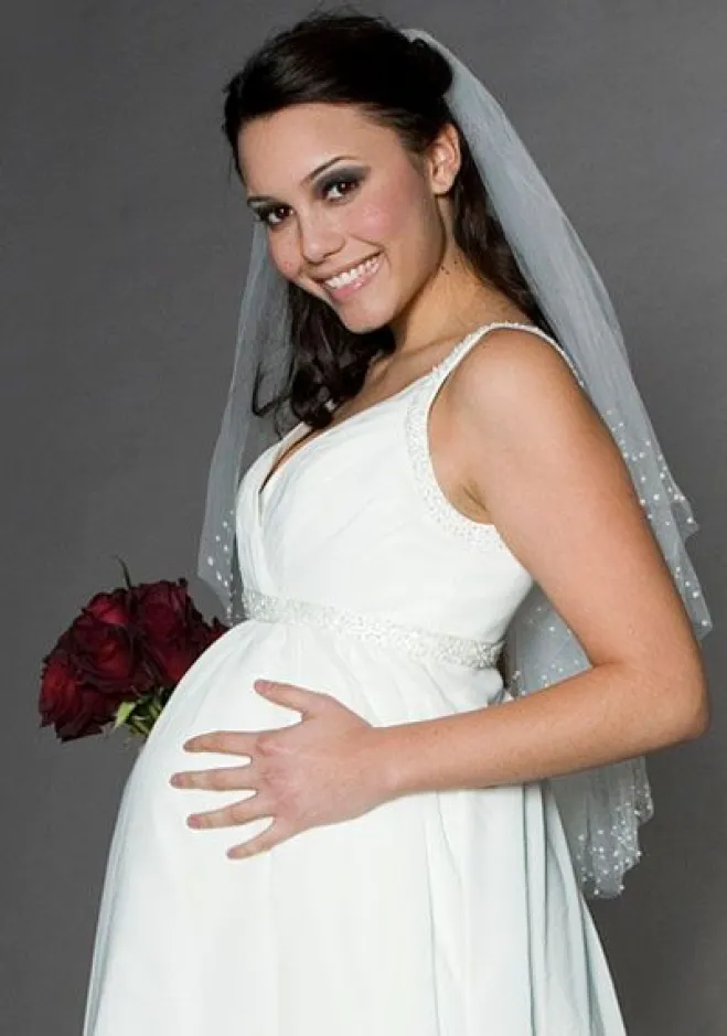 Mulher grávida usando um vestido de noiva. Ela sorri para a câmera e segura um buque de flores com uma das mãos.