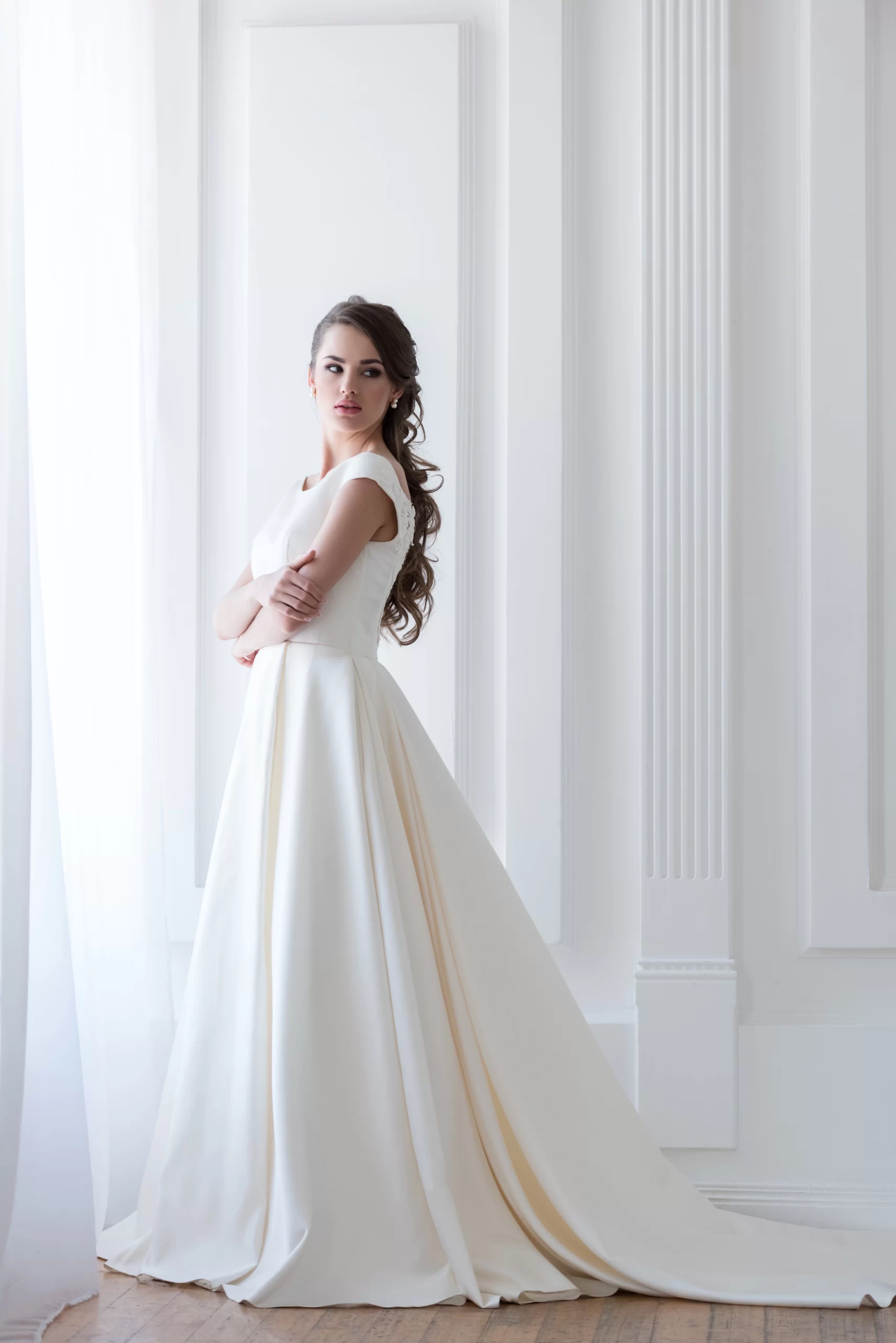 Mulher usando um vestido de noiva simples e branco com um corte tradicional. A mulher está de braços cruzados e está em uma posição virada de lado olhando para a câmera.