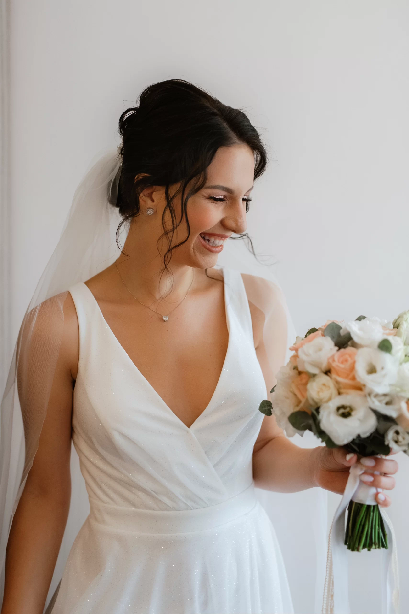 Mulher usando um vestido de noiva branco com um decote V. A foto da foco mostra a parte de cima do corpo da mulher. Ela está sorrindo e olhanldo para o buque que ela está segurando.