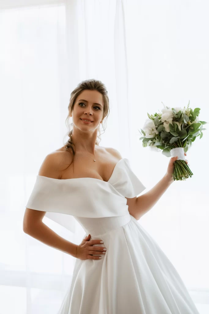 Mulher usando um vestido de noiva branco simples, ombro a ombro. Ela segura um buque de flores com uma das mãos e olha para o lado.