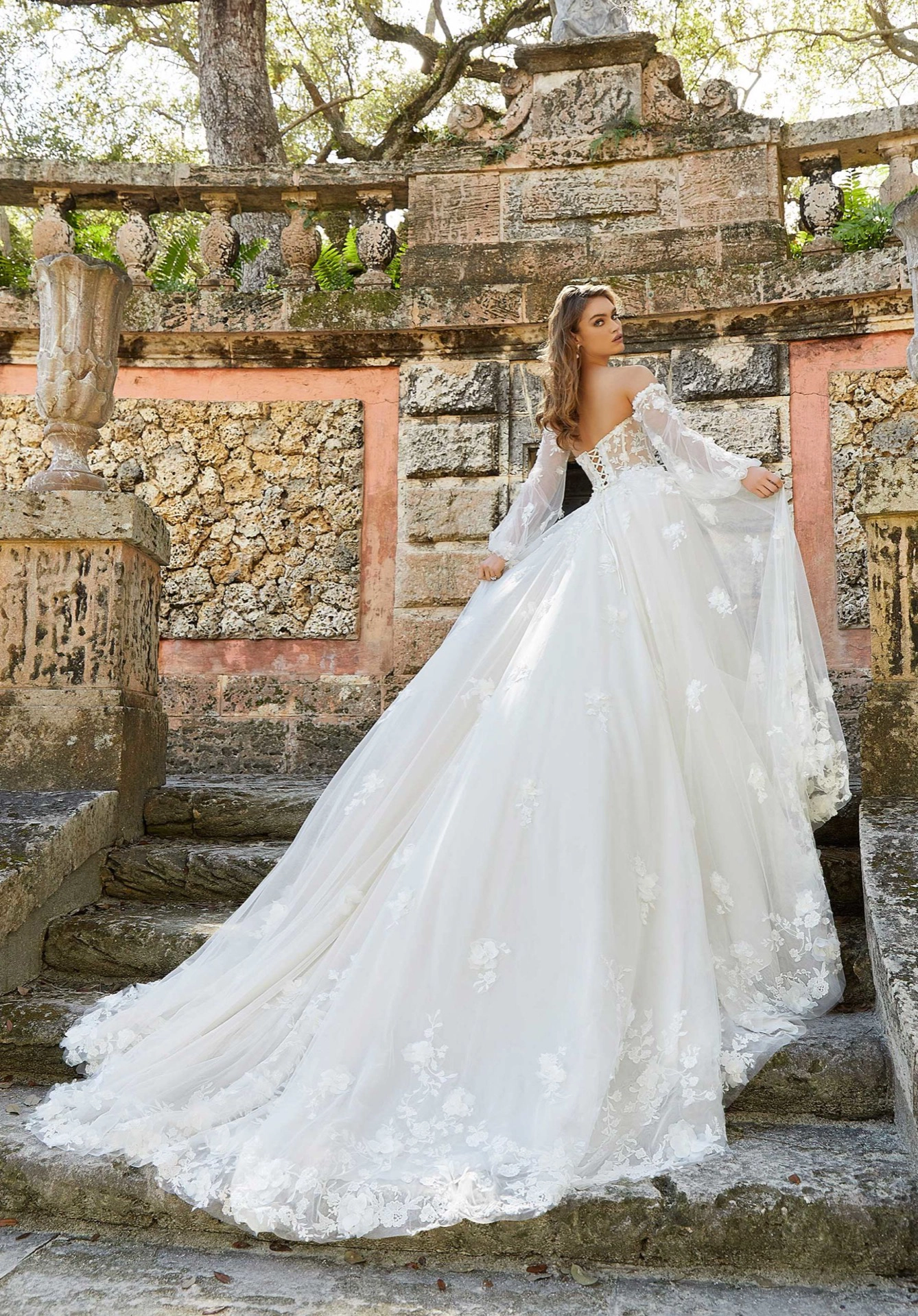 Mulher de cabelos castanhos claros usando um vestido de noiva branco de cauda longa. Ela está subindo uma escada de pedra olha para trás segurando as laterais do vestido.