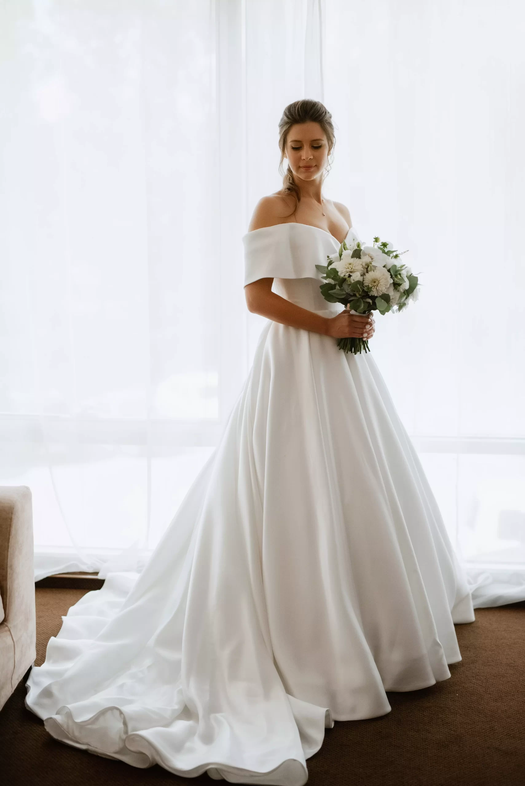 Mulher usando um vestido de noiva branco. Ela segura um buque de flores com ambas as mãos e olha levemente para baixo.