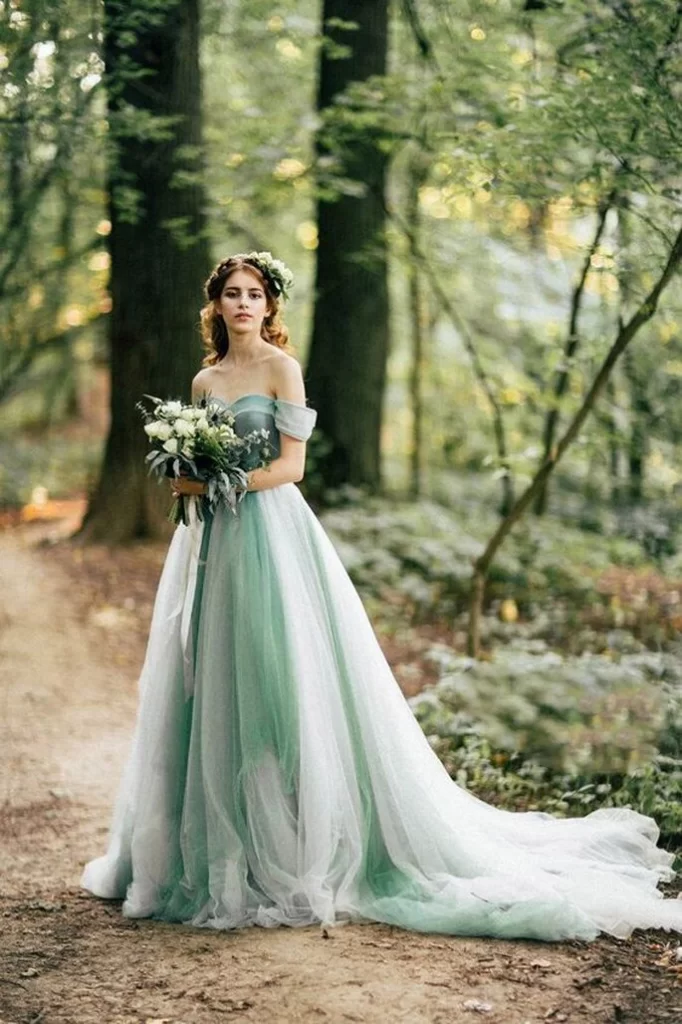 Mulher de cabelos castanhos em uma floresta usando um vestido de noiva verde. Ela segura um buque de flores brancas em ambas as mãos e olha para a câmera.